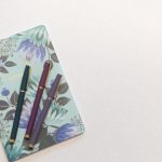 A notepad with floral prints in blue colors and three multi-colored pens on top
